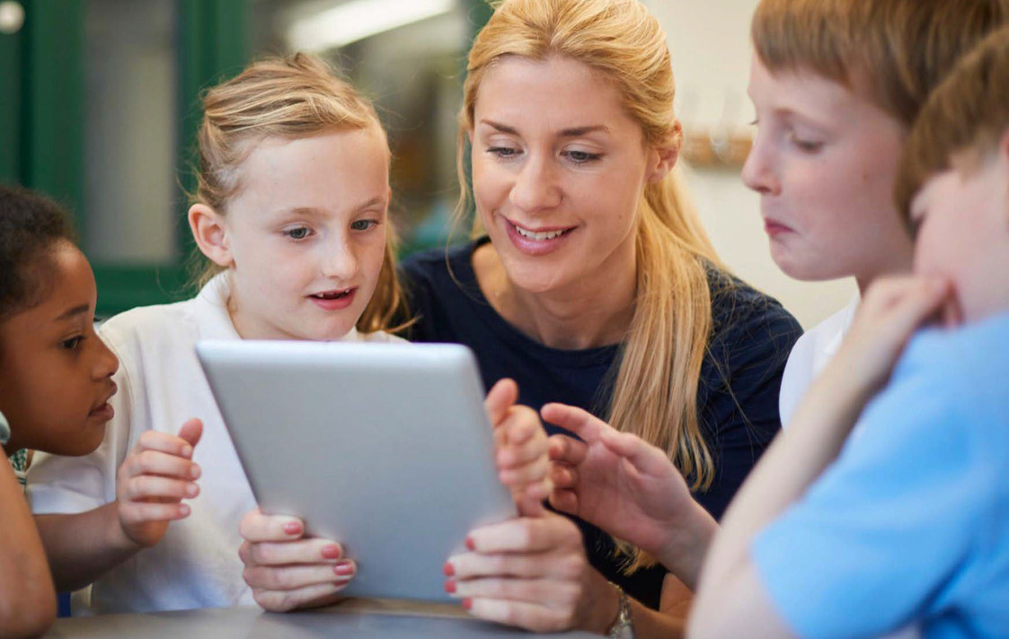 A teacher holding a tablet with children interacting with said tablet